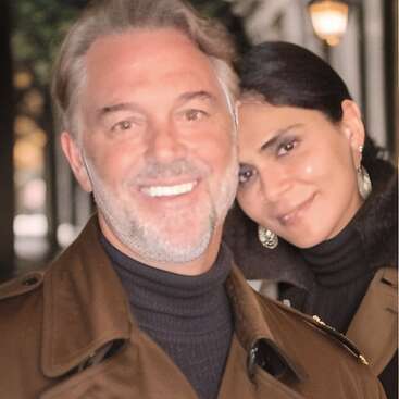 The image depicts a man and woman posing for a photo, with the woman\'s head resting on the man\'s shoulder, both wearing brown coats and black turtlenecks.