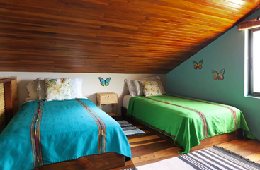 A imagem mostra um quarto com duas camas, cada uma com uma estrutura de madeira e um colchão branco, situado em um cômodo com piso de madeira e uma janela. As paredes são pintadas de branco.