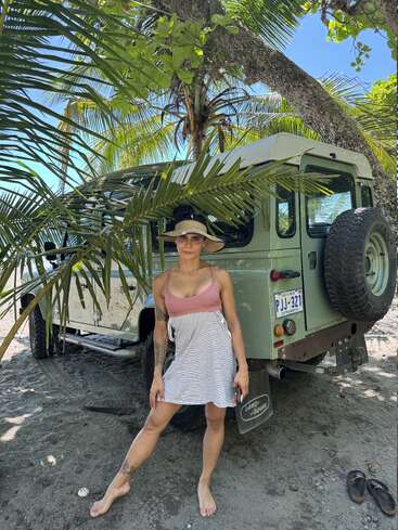 A imagem mostra uma mulher em frente a um Land Rover Defender verde em uma praia de areia, cercada por palmeiras e um céu azul claro.