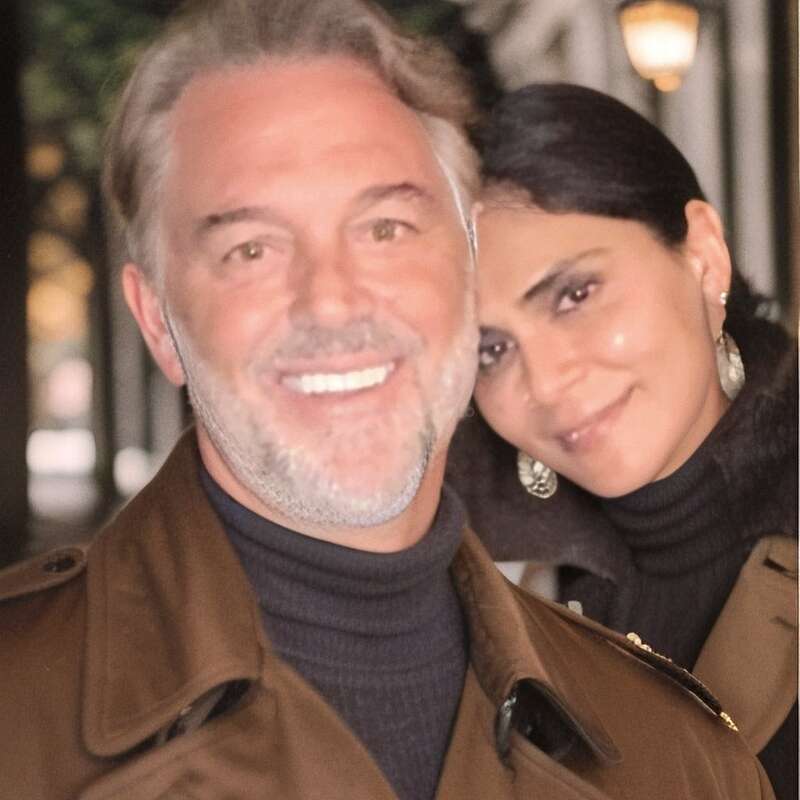 A imagem mostra um homem e uma mulher posando para uma foto, com a cabeça da mulher apoiada no ombro do homem, ambos vestindo casacos marrons e gola alta preta.