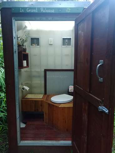Um banheiro rústico ao ar livre chamado "Le Grand Release" com interior de madeira, porta aberta, papel higiênico, pequena prateleira, plantas, duas aberturas e um ambiente natural sereno.
