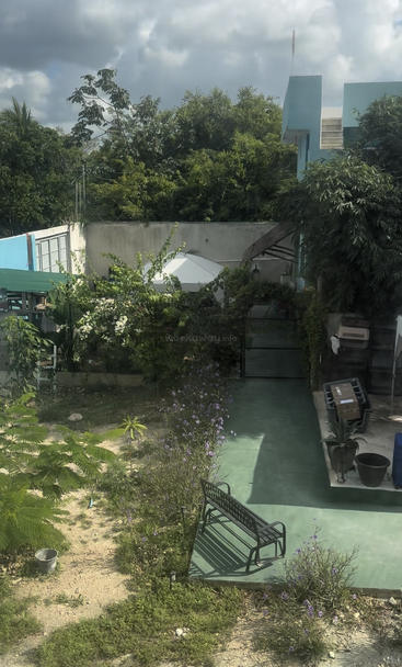 A peaceful garden with lush greenery, a black metal bench, potted plants, purple flowers, a green walkway, blue house, and partly cloudy sky overhead.