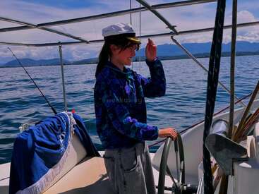 L'image représente une femme debout sur un bateau, tenant le volant de la main gauche, portant un chapeau de capitaine blanc et une chemise bleue à motifs.