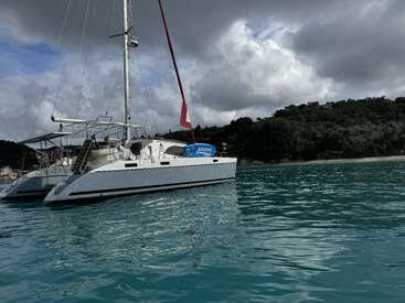 L'image représente un voilier catamaran blanc flottant sur des eaux calmes et bleues, avec un mât rouge et une enseigne bleue avec un texte blanc, le tout dans un ciel nuageux et sur un rivage boisé.