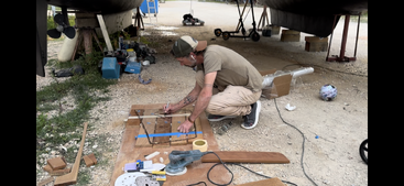 Un homme travaille sur un bateau dans une cour, entouré d'outils et de matériaux, avec un grand plan d'eau visible à l'arrière-plan. La scène se déroule sur un fond naturel.