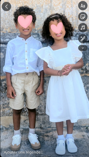 Duas crianças estão lado a lado, sorrindo para a câmera. O menino está com uma camisa branca e shorts bege; a menina está com um vestido branco. Os rostos estão cobertos por corações.