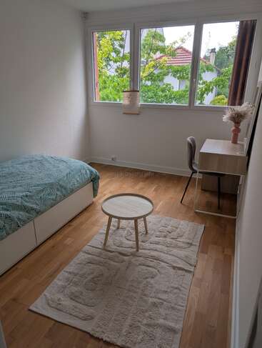 Este é um quarto bem iluminado e minimalista, com uma cama de solteiro, uma mesa de trabalho moderna, uma cadeira, uma mesinha de centro redonda, um tapete decorativo, uma janela grande e uma vista agradável para o exterior.
