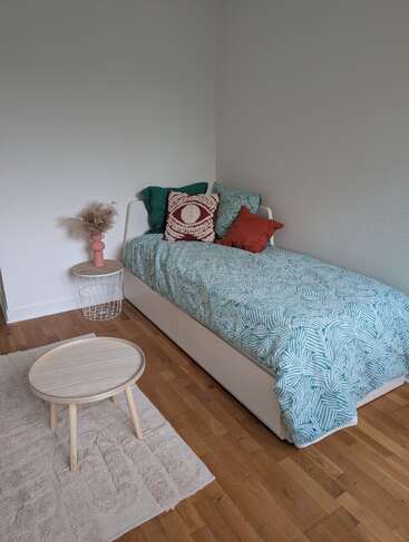 Um quarto minimalista tem uma cama de solteiro com roupa de cama azul-petróleo estampada e almofadas coloridas, uma mesinha redonda, um tapete texturizado e uma mesinha lateral decorativa com capim-dos-pampas.