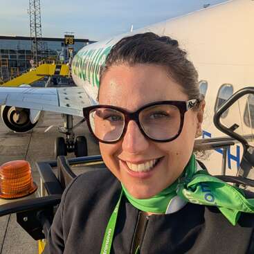 Uma mulher sorridente, de óculos e com um lenço verde, está em frente a um avião na pista, provavelmente uma funcionária do aeroporto, aproveitando o dia ensolarado.