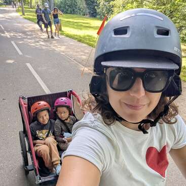 Uma mulher usando capacete e óculos escuros tira uma selfie enquanto anda de bicicleta. Duas crianças com capacetes estão sentadas em um carrinho de bicicleta atrás dela, numa ciclovia ensolarada.