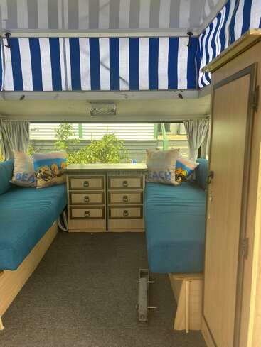 This image shows the cozy interior of a camper with blue-striped canopy, blue cushioned benches, beach-themed pillows, wooden storage drawers, and a small window.