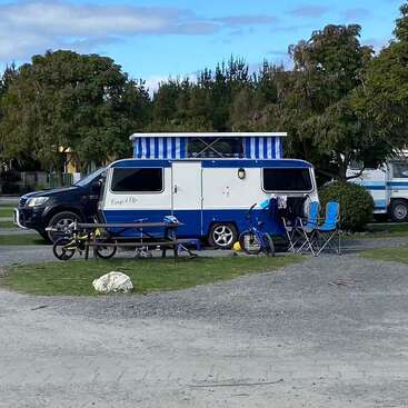 A blue and white camper van is parked at a campsite with foldable chairs, bicycles, picnic table, and trees in the background on a partly cloudy day.