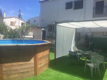 La imagen muestra un patio trasero con piscina, césped artificial y muebles de exterior, sobre un fondo de casas y árboles en un día soleado.