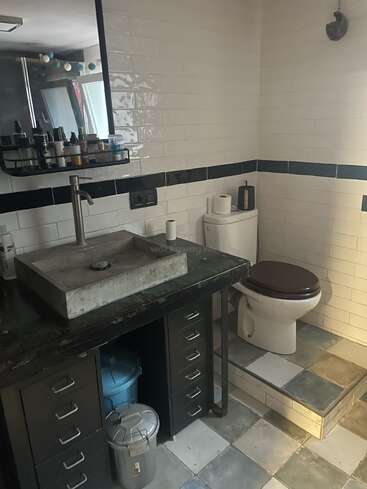 Este cuarto de baño cuenta con un inodoro elevado, un lavabo de estilo industrial con cajones, un espejo grande, artículos de aseo, papeleras, baldosas de metro blancas y baldosas estampadas en el suelo, que crean un aspecto ecléctico.