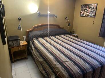 Un acogedor dormitorio con una cama grande, ropa de cama a rayas, dos mesillas de noche, lámparas de lectura en la pared, fotos en la pared y una pequeña ventana con cortinas.