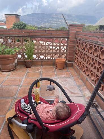 Un bebé en una mecedora roja está sentado en un patio embaldosado, rodeado de macetas y paredes de ladrillo, con las montañas y el cielo nublado de fondo.