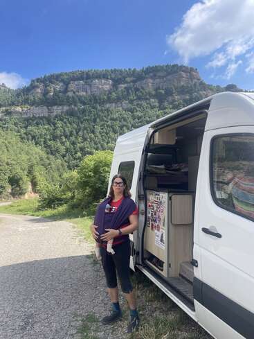 Una mujer, de pie junto a una furgoneta blanca, lleva a un bebé en un fular portabebés. Detrás de ellos hay frondosos bosques verdes y acantilados rocosos bajo un cielo azul brillante.