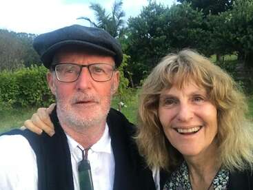 La imagen muestra a un hombre y una mujer posando para un selfie en un jardín, con el hombre con gorra plana y chaleco, y la mujer con el pelo rubio.
