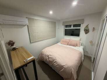 Este quarto aconchegante tem uma cama bem arrumada com roupa de cama em rosa suave, uma mesa de madeira, quadros na parede, ar-condicionado, persianas bege e uma decoração minimalista.