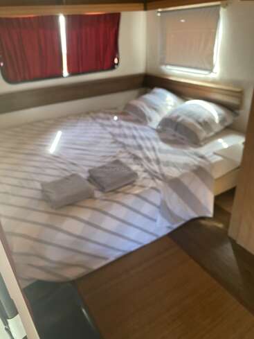 This image shows a cozy bedroom with a neatly made double bed, striped bedding, two pillows, folded towels, wooden floor, and windows with red and white curtains.