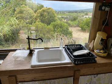 A cozy kitchen sink with a rustic wooden counter overlooks a lush green landscape through a wide window, featuring clean dishes, soap, and a yellow coffee maker.