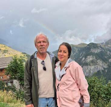A man and woman stand together outdoors in mountainous scenery, smiling, with a faint rainbow in the cloudy sky behind them. Greenery and wooden building nearby.