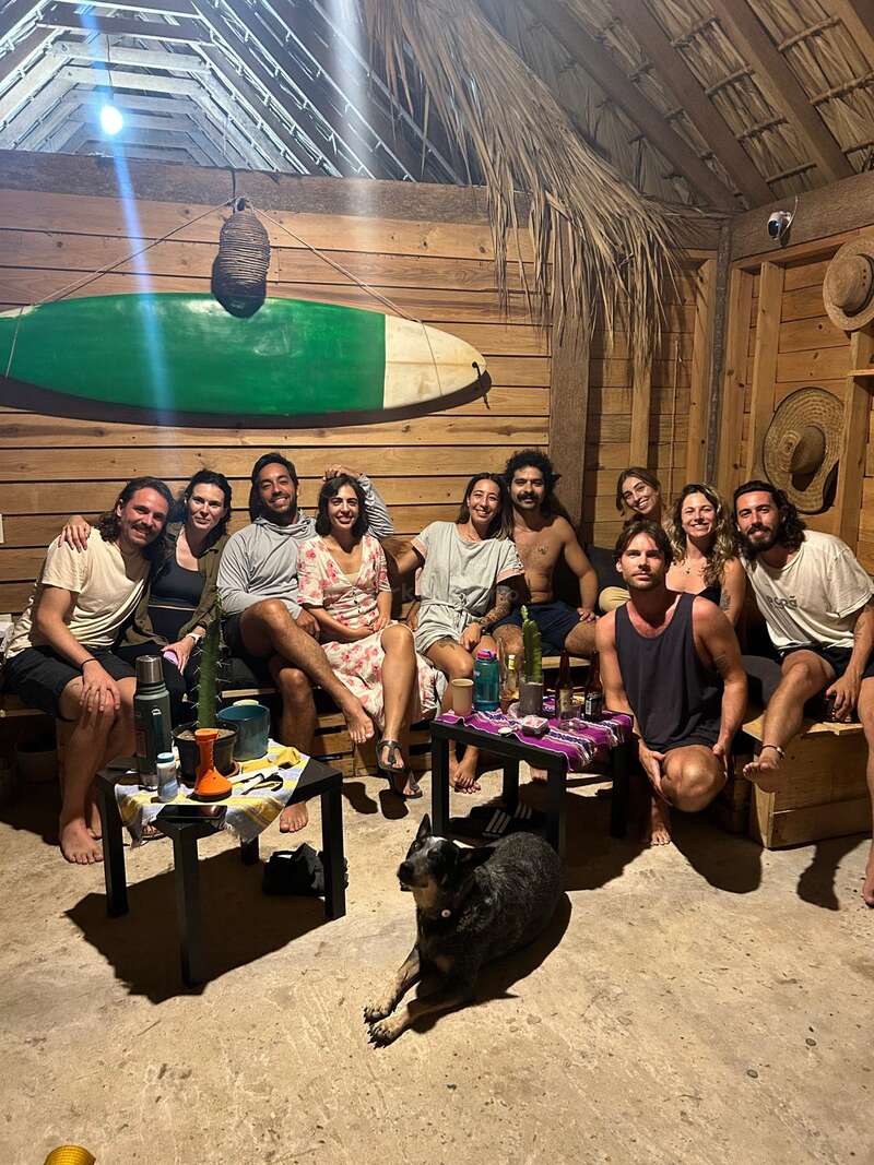 A imagem mostra um grupo de nove pessoas e um cachorro em uma sala aconchegante com paredes de madeira, com uma prancha de surfe na parede e um cachorro deitado no chão.