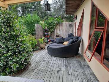 A cozy outdoor patio features a round wicker lounge chair with cushions, surrounded by lush greenery, potted plants, wooden fencing, and large windows with red frames.