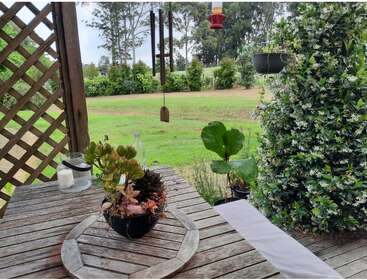 A rustic wooden table with potted succulents sits on a porch overlooking a lush, green garden, hanging plants, wind chimes, and distant trees under a cloudy sky.