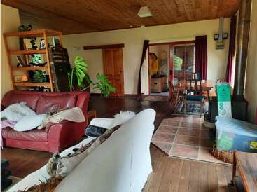 Cozy living room with wooden ceiling, red and white sofas, bookshelf, green plants, dining area, large windows with curtains, and assorted household items neatly arranged.