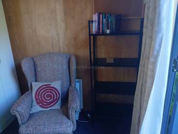 A cozy reading corner with a patterned armchair, spiral pillow, empty black bookshelf with a few books on top, wooden panel walls, and natural light from a window.