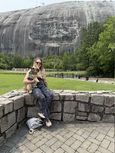 Eine Frau mit Sonnenbrille sitzt auf einer Steinmauer und hält einen kleinen Hund im Arm. Hinter ihr erhebt sich ein großer Felsberg mit einem gemeißelten Denkmal und Bäumen.
