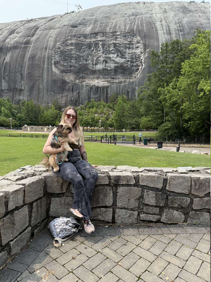 Eine Frau mit Sonnenbrille sitzt auf einer Steinmauer und hält einen kleinen Hund im Arm. Hinter ihr erhebt sich ein großer Felsberg mit einem gemeißelten Denkmal und Bäumen.