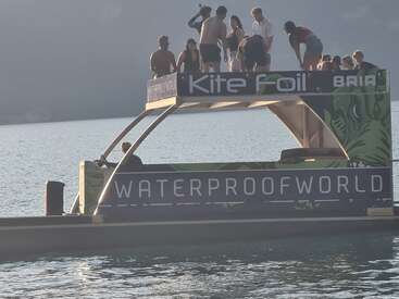 Um grupo de pessoas está em uma plataforma flutuante com as inscrições “Kite Foil” e “WATERPROOFWORLD” em um lago calmo, curtindo um dia ensolarado.