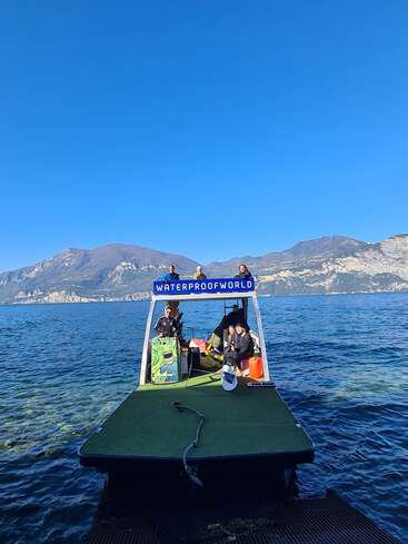 Um grupo de pessoas em uma plataforma flutuante com a inscrição “WATERPROOF WORLD”, cercada por águas azuis, montanhas ao fundo, sob um céu azul claro e brilhante.