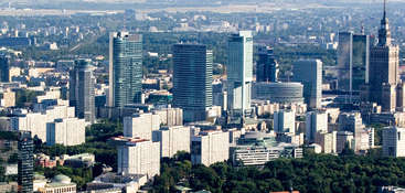 A imagem mostra uma paisagem urbana com um edifício proeminente, provavelmente um arranha-céu, situado no centro, cercado por outros edifícios e árvores, com um céu claro acima.
