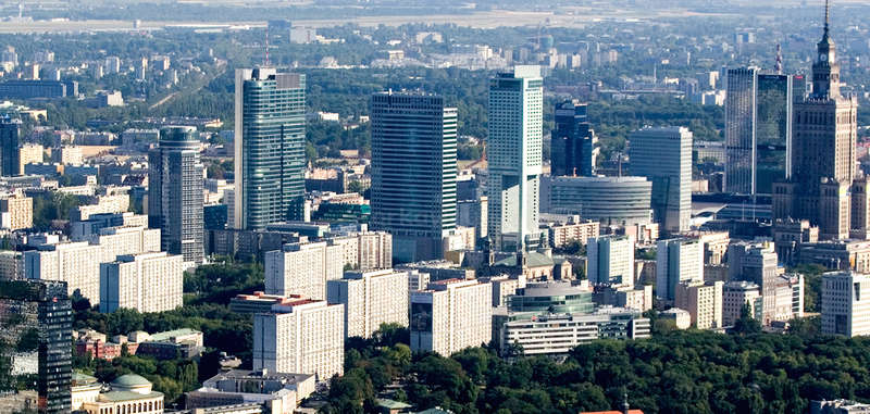 The image depicts a cityscape with a prominent building, likely a skyscraper, situated in the center, surrounded by other buildings and trees, with a clear sky above.