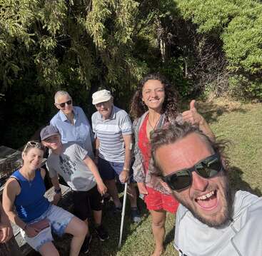 Six people pose happily outdoors on a sunny day, surrounded by trees and grass. They smile at the camera, enjoying a cheerful moment together in nature.