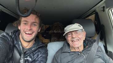 A young man and an elderly man smile happily while sitting in the front seats of a vehicle. Both wear jackets; a dog sits in the back.