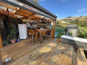 This image shows a rustic outdoor kitchen with wooden floors, potted plants, a dining table, refrigerator, hanging cookware, and scenic mountain views under a clear sky.