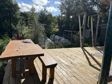 A rustic wooden deck with a table and bench overlooks a lush garden with trees, greenhouses, and swings under a bright, clear blue sky.