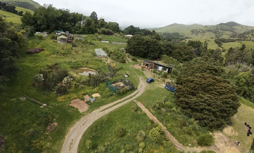 Eine ländliche Landschaft mit einem gewundenen Feldweg, grünen Feldern, Gärten, einem kleinen Teich, verstreuten Bäumen, einem Haus, geparkten Autos und fernen Hügeln. Eine friedliche Szene auf dem Land.