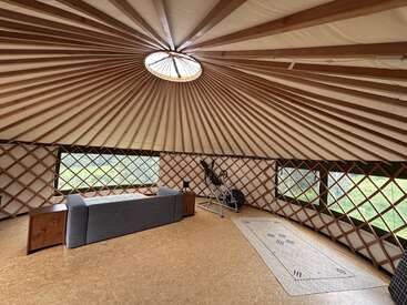 Dieses Bild zeigt das Innere einer geräumigen Jurte mit Holzgitterwänden, einem runden Dachfenster, einem grauen Sofa, Fitnessgeräten und minimalistischen Holzmöbeln.