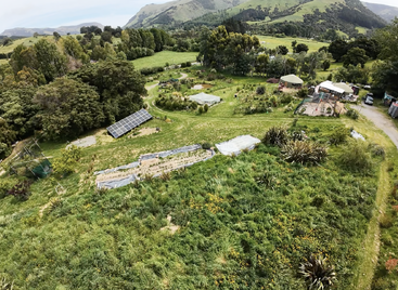 Eine üppige grüne Landschaft mit Sonnenkollektoren, Gemüsegärten, Jurten und einer kleinen Bauernhofsiedlung, umgeben von Bäumen, Hügeln und einer lebendigen natürlichen Vegetation. Eine friedliche, ländliche Umgebung.