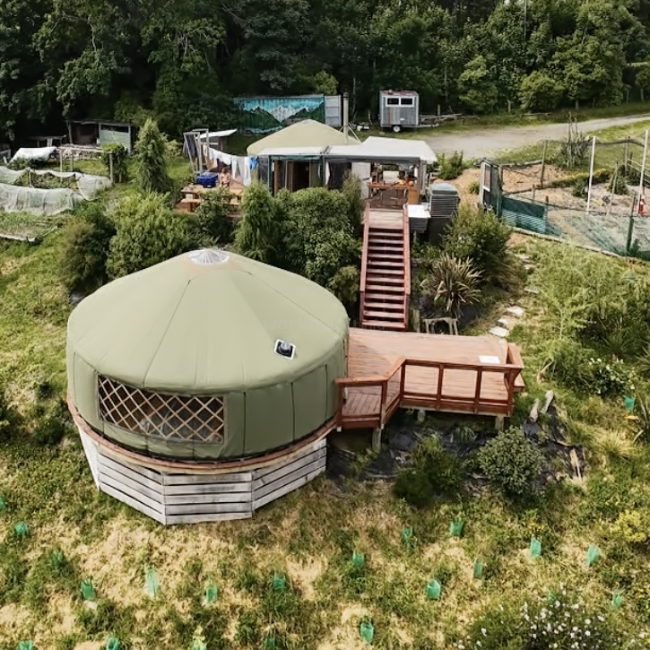 Eine grüne Jurte steht auf einem Hügel, umgeben von üppiger Vegetation, Holzdecks, Treppen, Gärten und Bäumen, die sich harmonisch in die ländliche Landschaft einfügen.