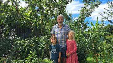 Um homem mais velho e duas crianças pequenas estão juntos em um jardim verde e exuberante. Árvores altas e várias plantas os cercam sob um céu azul brilhante.