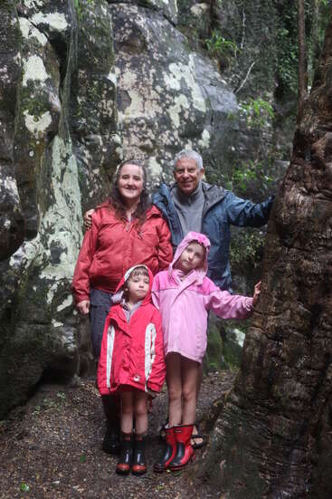 Uma família de quatro pessoas, vestida com jaquetas e botas de chuva, está reunida em uma floresta exuberante e rochosa. Eles parecem felizes, explorando a natureza em um dia chuvoso.