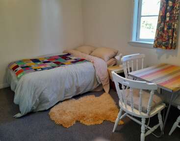 Esse quarto aconchegante tem uma cama de solteiro com cobertor colorido de retalhos, travesseiros bege macios, um tapete fofo, uma mesa pequena, duas cadeiras, janela clara e cortinas.