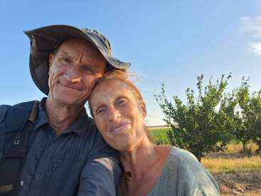 A smiling couple takes a selfie outdoors on a sunny day, standing closely together with trees and a blue sky in the background, radiating happiness and warmth.