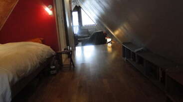 This cozy attic room features wooden floors, a bed with white bedding, angled walls, a small side table, bookshelves, and a distant armchair by a window.